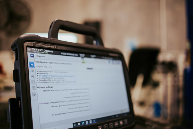 Close-up of a diagnostic tablet with the Advanced Diagnostics vehicle application open, displaying progress and optional settings. The background is blurred, indicating an indoor workshop.